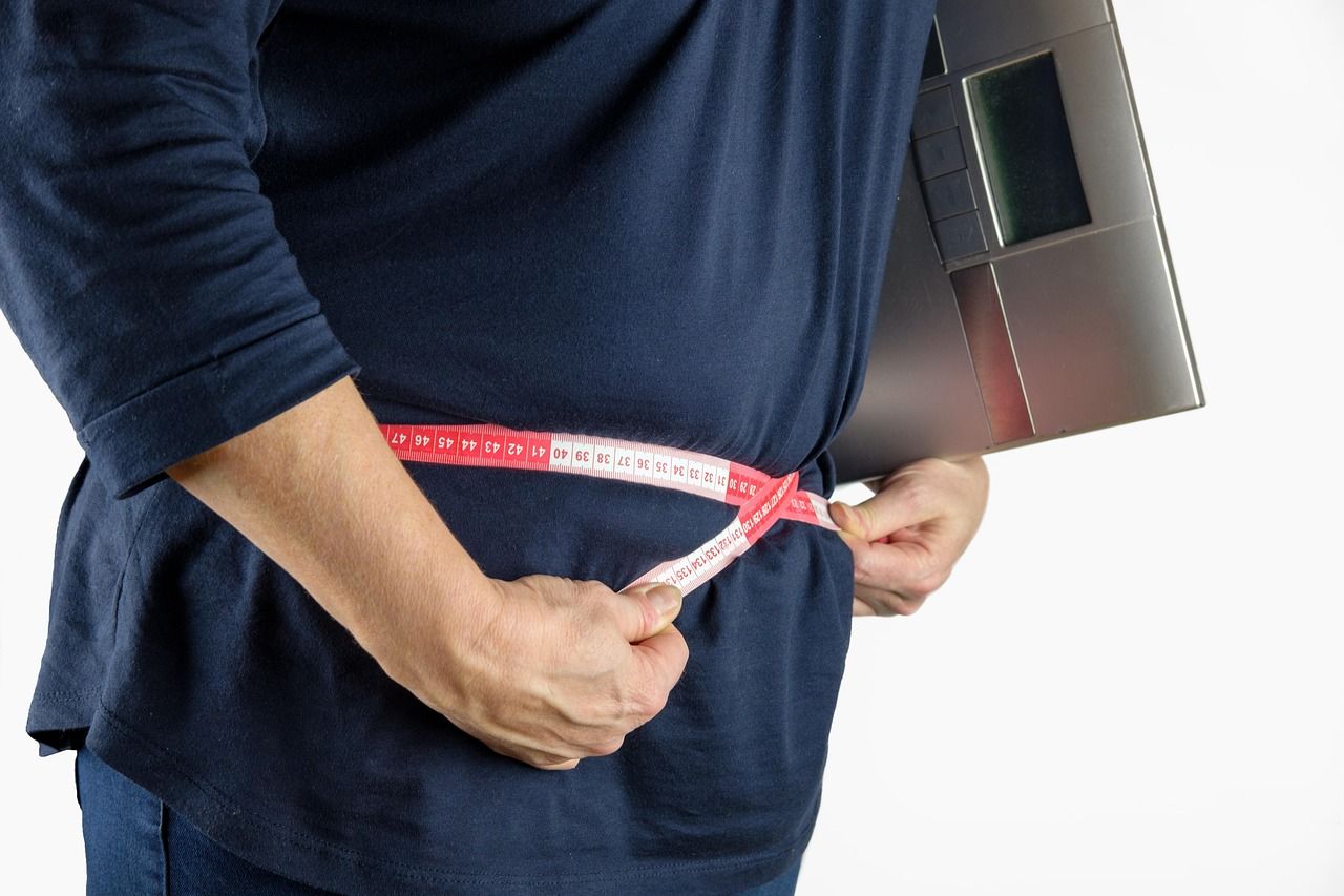Žena s viškom kilograma u tamnoplavoj majici krojačkim metrom mjeri opseg struka, a pod rukom drži osobnu vagu